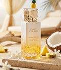 Der weiß-goldene Glasflakon von Gulf Orchid Tahiti Colada mit kleiner Ananas auf dem Deckel, tropisch präsentiert am Strand mit frischer Ananas und Kokosnuss.